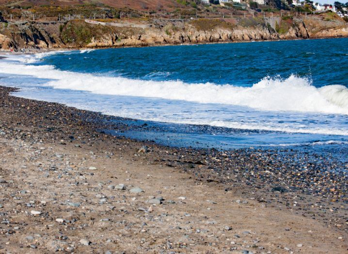 Image of a beach with rocks and sand on the shoreline and white foam visible on the waves.