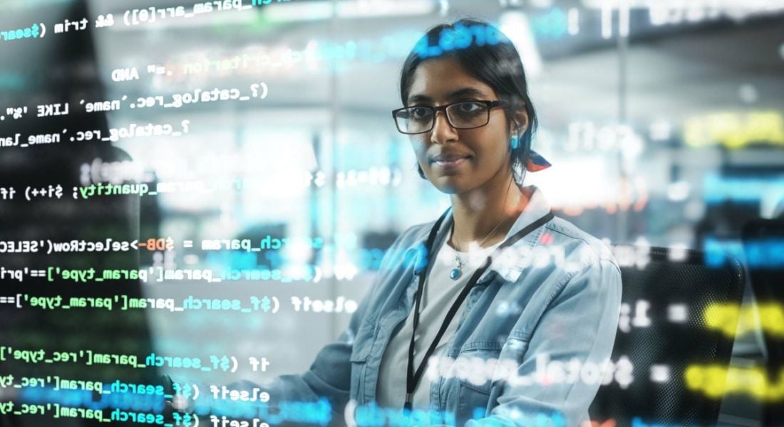 Lines of code reflected on a woman's face as she stands in front of a computer screen in an office environment.