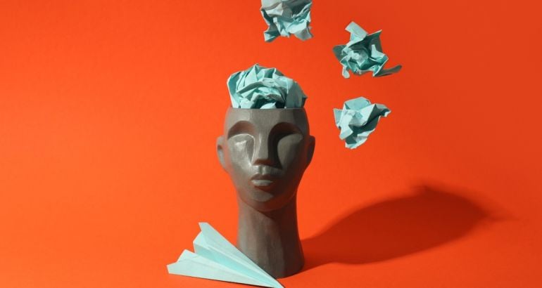 A grey head statue with a green paper plane and green balled up pieces of paper coming out of its head. The background is orange.