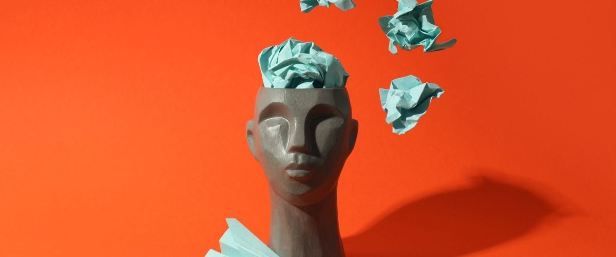 A grey head statue with a green paper plane and green balled up pieces of paper coming out of its head. The background is orange.