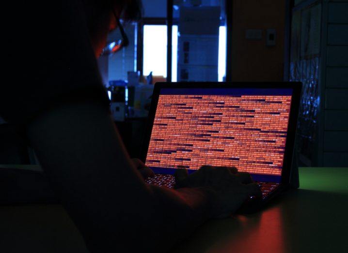 A person in a dark room typing on a laptop with red code on the screen. Used to illustrate the concept of ransomware.