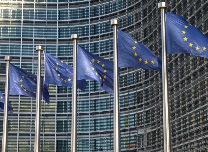 EU flags on poles flying against a big glass fronted building.