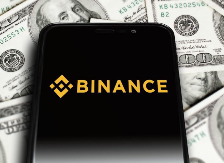 The Binance logo on the screen of a smartphone, which is laying on top of multiple US dollar bills.