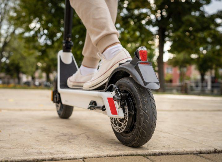 A person's legs on an e-scooter, with the back wheel of the vehicle being close and the front being in the background. There are trees in the background.