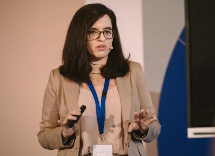 A woman stands on stage speaking at an event. She is Aikaterini Liakopoulou.