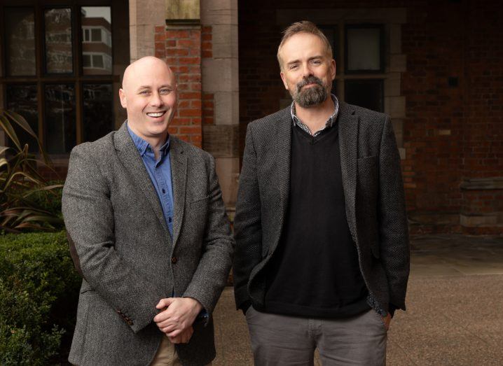 Two men stand next to each other in the outdoors and smile.