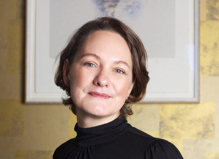 A headshot of Nicola Baker from Biologit. She has short brown hair and wears a black turtleneck top. She stands in front of a yellow wallpapered wall with a framed picture of a flower drawing.