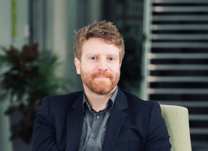 A man in a black suit sitting down indoors, with a plant and stairs visible in the background. He is Brendan Carroll, the founder and CEO of EpiSensor.