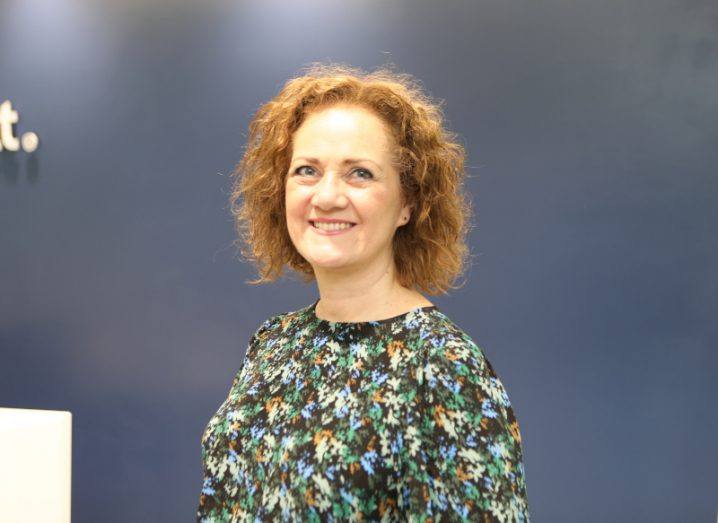 A woman with a patterned top smiles at the camera in front of a dark grey wall. She is Claudia Carr, a partner at BearingPoint.