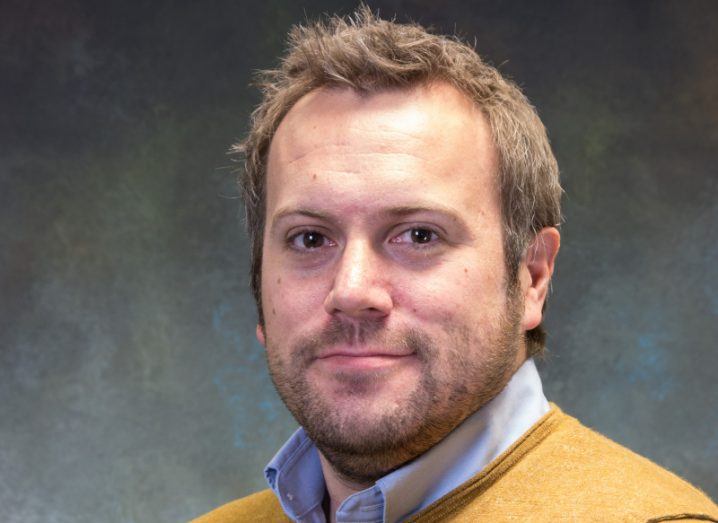 A man wearing a yellow jumper over a white shirt smiles at the camera in front of a dark grey wall. He is Dave Adamson, chief technology officer at Espria.