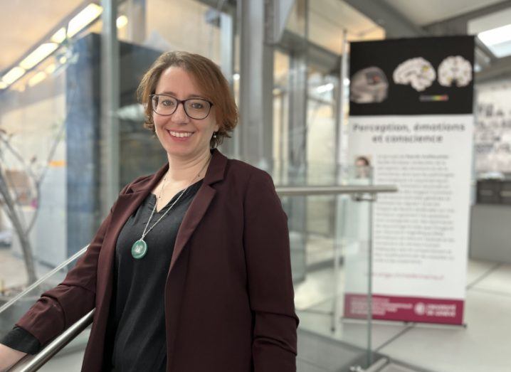 Kimberly Doell wears a wine-coloured blazer and black shirt and stands with one arm rested on a banister in a large glass building.