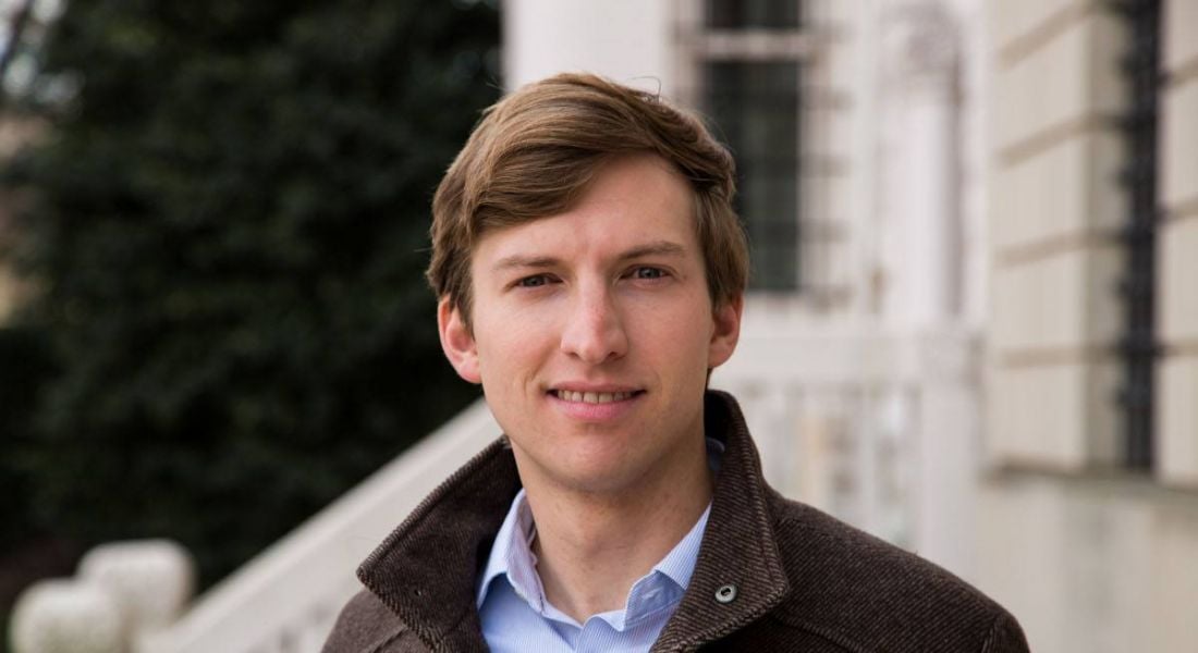 A headshot of Prof Chris Miller wearing a heavy brown jacket and standing outside in front of a grand building entrance.