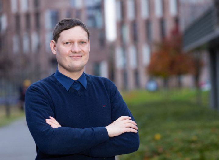 Andrei Ermakov stands with his arms folded, wearing a navy blue jumper and blue shirt. outside with grass and a path behind him and a large building in the background.