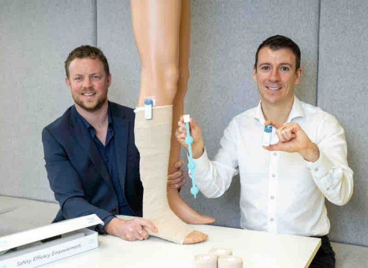 Two men stand at a table with a dummy leg bandaged. One man is holding a device with another attached to the bandage.