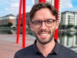 A man wearing a grey T-shirt smiles at the camera with his arms folded in an office setting. He is Robert McCullough, a senior solutions architect at Liberty IT.