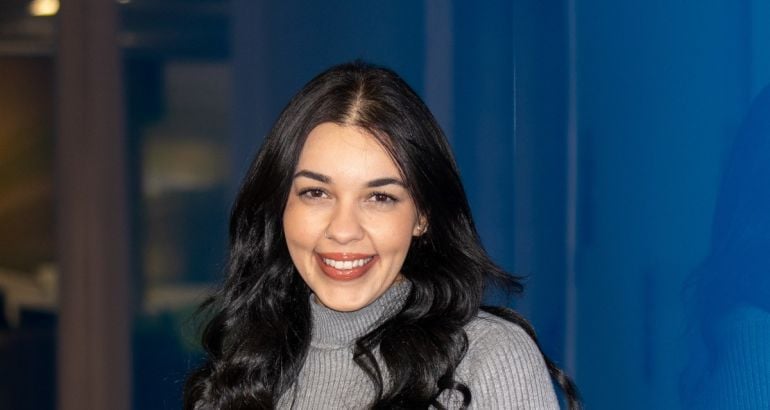 A young woman with long dark hair against a blue background wall smiles at the camera. She is Geovana Lore of Intel.