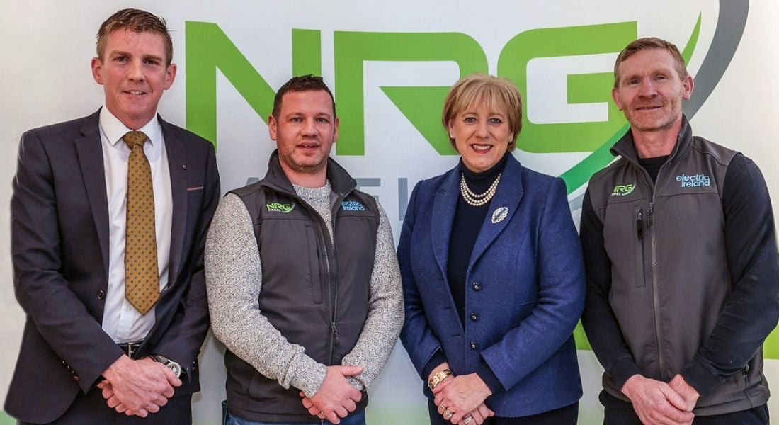 Three people from NRG Panel and Minister Heather Humphreys, TD standing in front of a sign for NRG Panel.