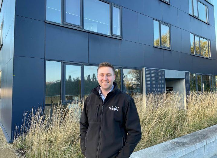 A man stands in front of a building with his hands in his pockets. He is smiling at the camera.