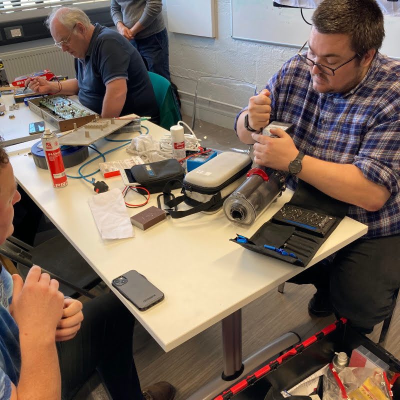 Two men sitting at a table working on electrical equipment.