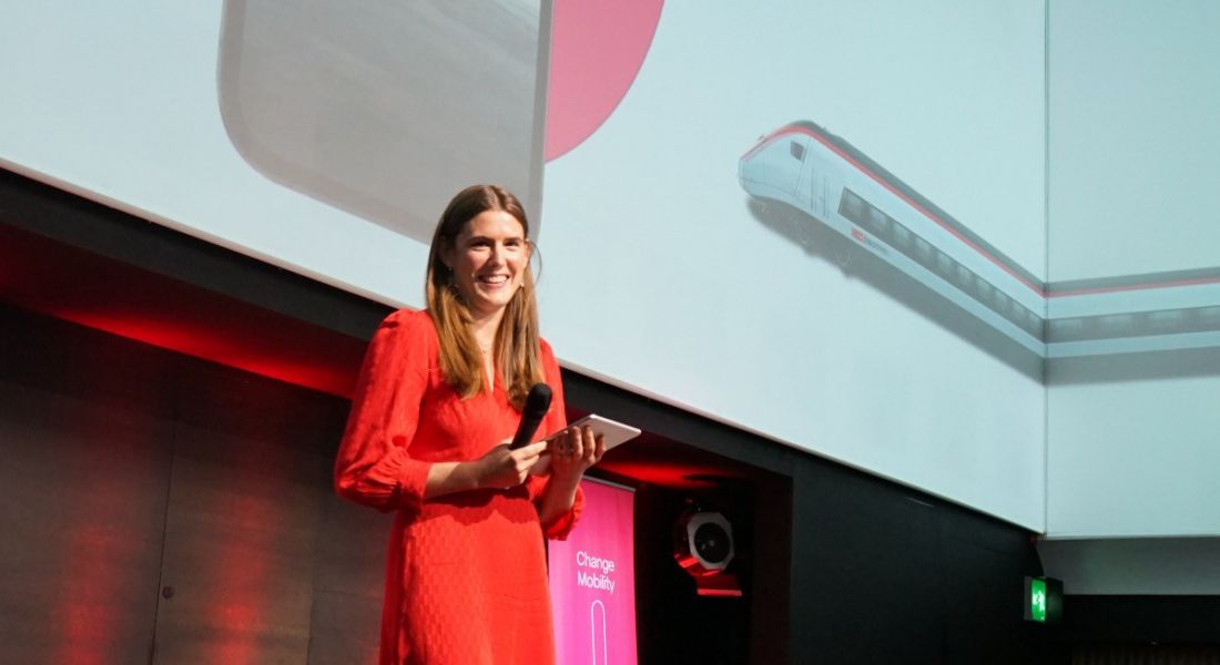 A woman in a red dress stands on stage with a screen behind her showing a train. She is Judith Häberli and she’s holding a microphone.