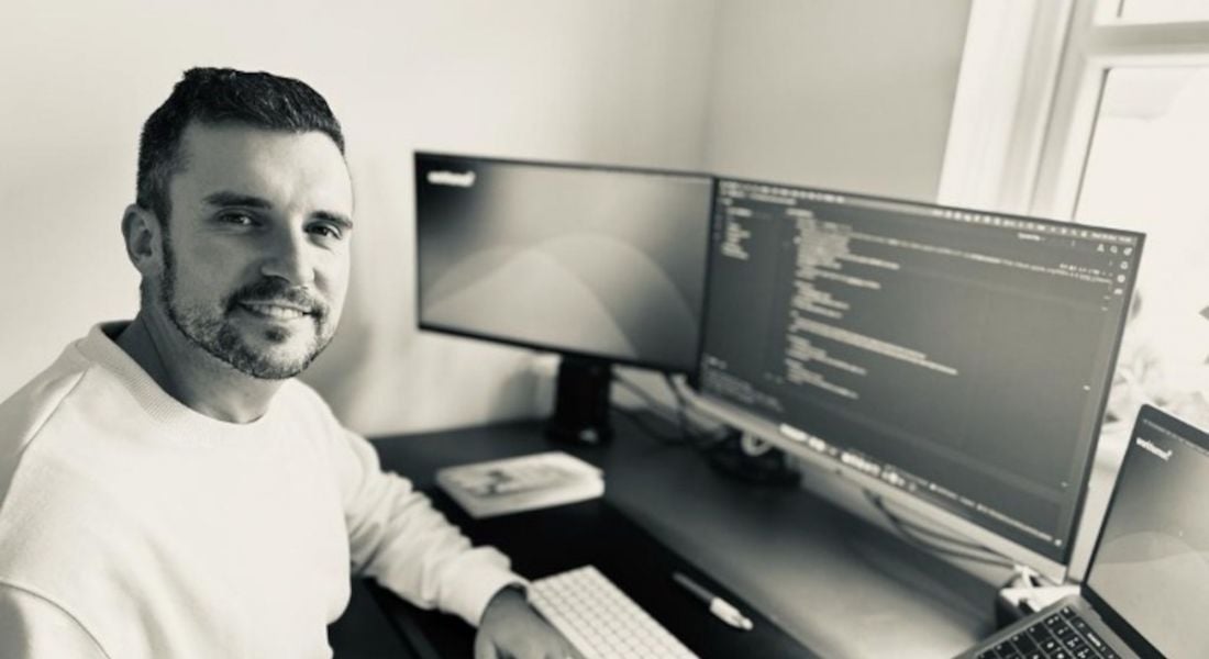 A man wearing a light-coloured jumper sits at a work desk and smiles to the camera. On his desk are two computer monitors, with one of the screens displaying lines of code. He is Keith Tuffy, a principal engineer at Workhuman.