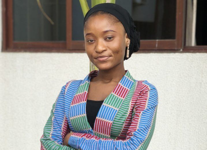 A young woman smiles at the camera with her arms folded. She stands outside a window and wears a stripey top.