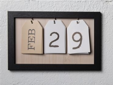 A picture frame with three tags hanging on it sits on a white wall. The first tag has the word February on it, while the other two tags spell out the number 29.