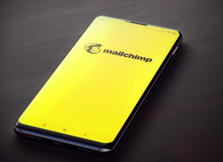 A phone with Mailchimp software is on the table. The yellow Mailchimp branding and the monkey logo are clearly visible on the phone screen.