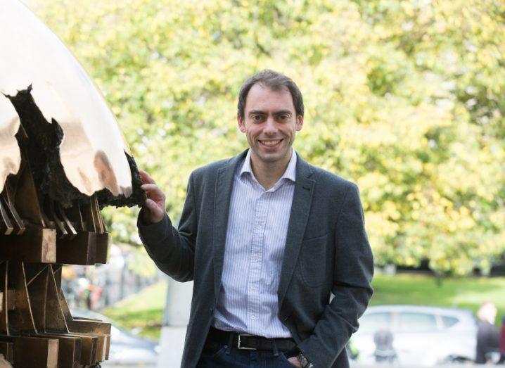 A man in a grey suit standing next to a statue, with trees in the background. He is Professor Marco Ruffini.