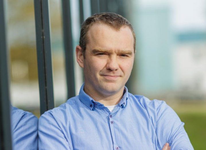 A headshot of Mark Pugh wearing a blue shirt, leaning against the side of a reflective building.