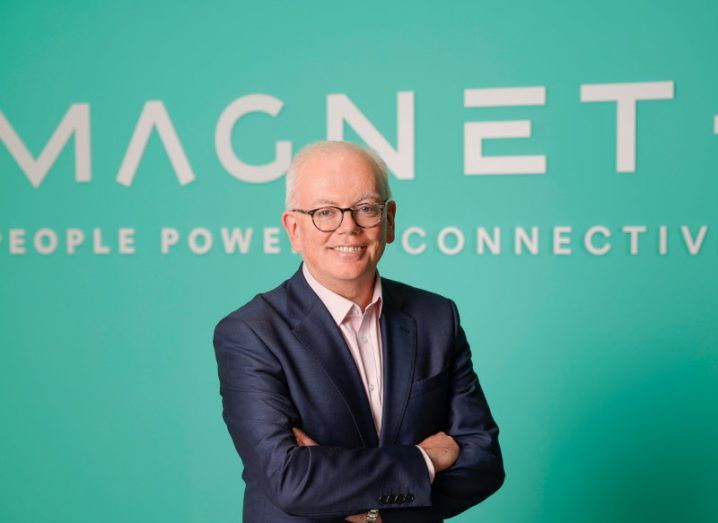 Patrick Masterson wearing a black suit and standing in front of a green wall that has the Magnet Plus logo on it.