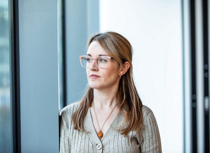 Jacqui O'Connor stand in front of floor to ceiling windows , looking to her right. She wears glasses and has long brown hair.