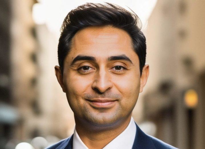 A close-up headshot of a man in a suit and white shirt, against a blurred urban background.