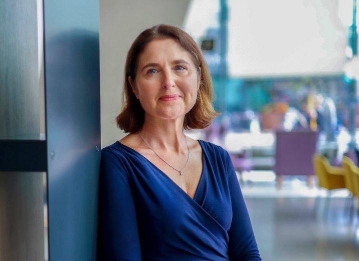 A woman in a blue outfit leaning against a grey wall. She is Rachael Ingle of Aon Ireland.