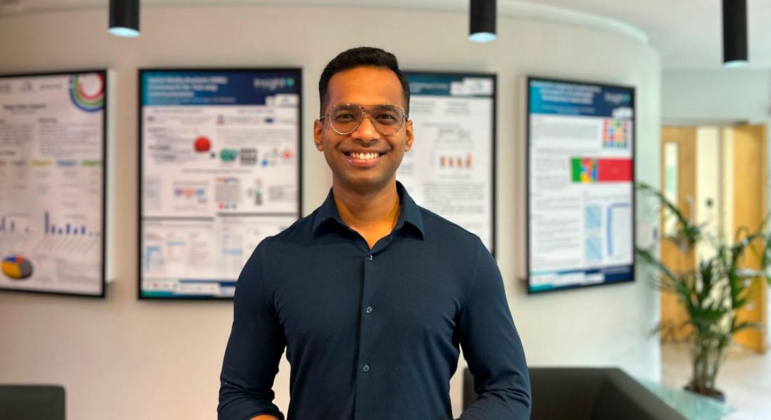 A man wearing a navy-coloured shirt and glasses smiles at the camera in front of a wall that holds various framed charts. He is Rajdeep Sarkar, a research scientist at Yahoo.