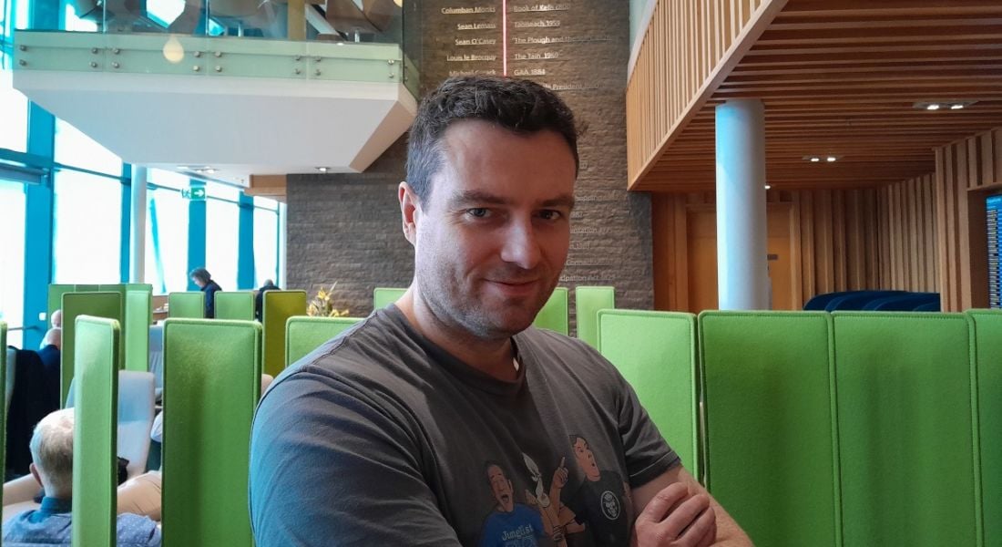 A man wearing a grey T-shirt smiles at the camera with his arms folded in an office setting. He is Robert McCullough, a senior solutions architect at Liberty IT.