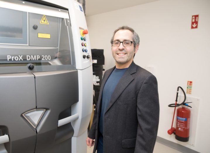 A photo of Dr Rocco Lupoi next to a large machine. He wears a t-shirt and dark blazer and smiles at the camera.