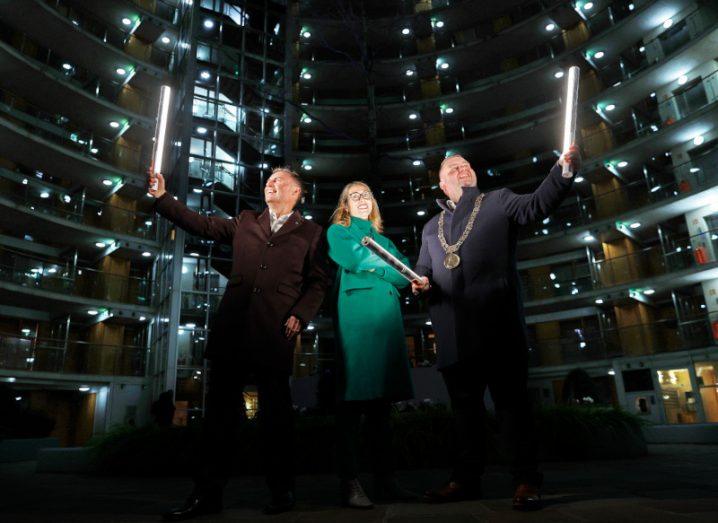 A woman wearing bright green stands between two men who are holding up light strips. The background is dark with spots of light.