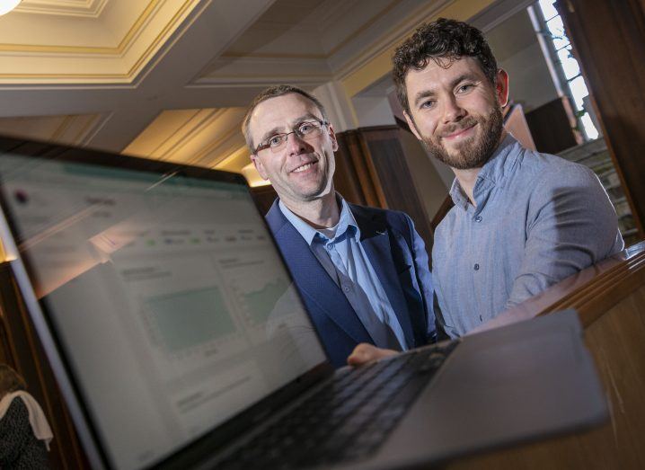 Two men stand and smile at the camera with a laptop in the foreground showing the Lua Health app.