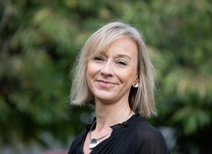 A headshot of a woman with a blonde bob, wearing a black shirt and a necklace, who smiles at the camera in front of blurry greenery outside.