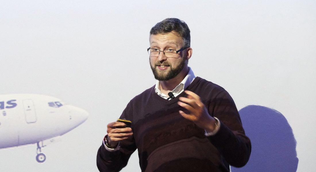 A man wearing glasses stands in front of a large screen, speaking at an event. He is Darktrace’s Toby Lewis.