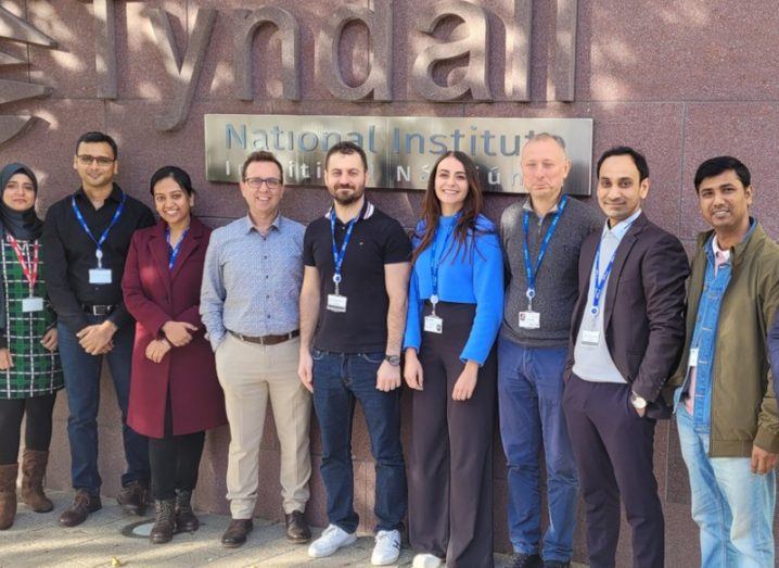 A group of men and women standing in front of a brown building with the Tyndall National Institute sign on the building wall.