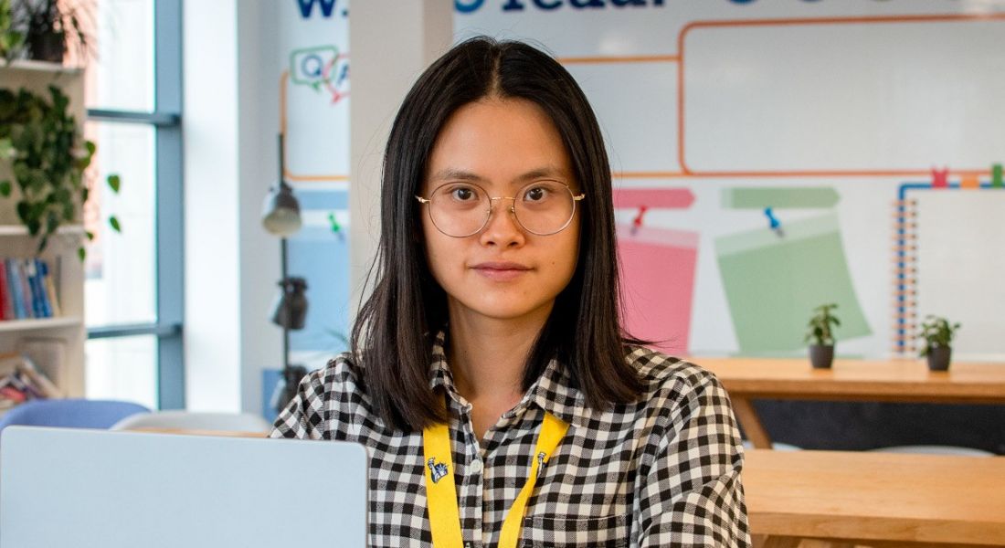 A woman wearing glasses and a chequered shirt smiles at the camera from behind a laptop. She is Wanqi Chen, an associate software engineer at Liberty IT.