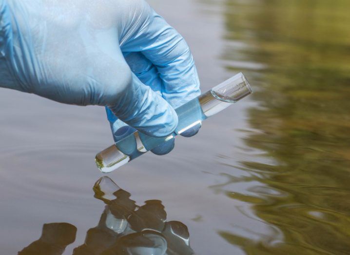 A blue-gloved hand is seen holding a test tube with water above an outdoor body of water to symbolise testing in the environment for PFAS.