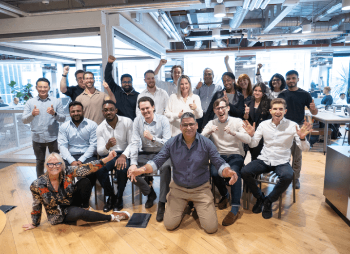 A group of men and women standing and sitting in a group in an office, with a wooden floor under them. They are the London team of Alt21.