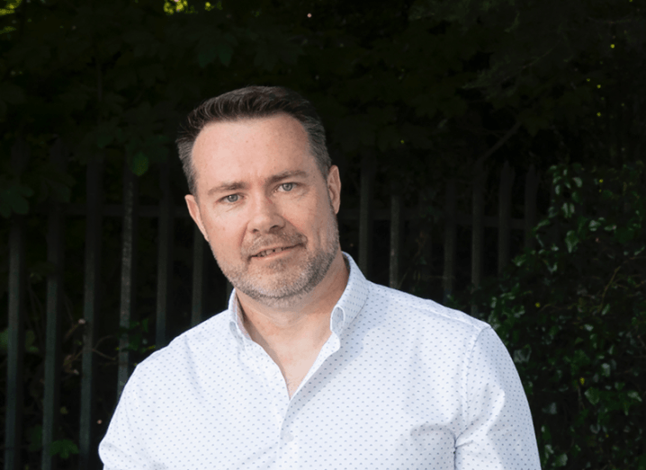 A man in a white shirt with a fence and green leaves behind him. He is a co-founder of Erapid.